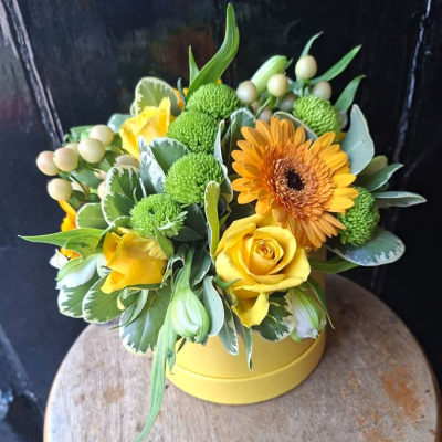 Sunshine Meadow Hat Box - A cheerful burst of sunshine in a bright yellow hat box, hand-arranged with golden roses, a sunny gerbera, lime-green chrysanthemums and ivory hypericum berries, finished with variegated foliage. A joyful gift to brighten any day.