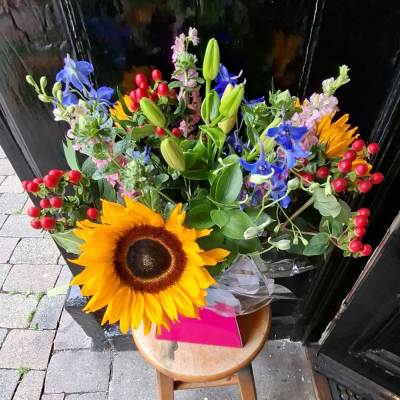 Lakeland Sunflower Bouquet - A striking statement hand-tie led by bold sunflowers, vivid blue delphinium, pink stocks, lily buds and bright red hypericum berries. A big, joyful gift that makes an impression.