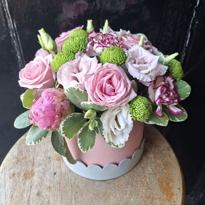 Blush Petals Hat Box - A pretty pink scalloped hat box filled with soft pink roses, ruffled lisianthus, green button chrysanthemums and striped carnations, framed with variegated foliage. Delicate and romantic.
